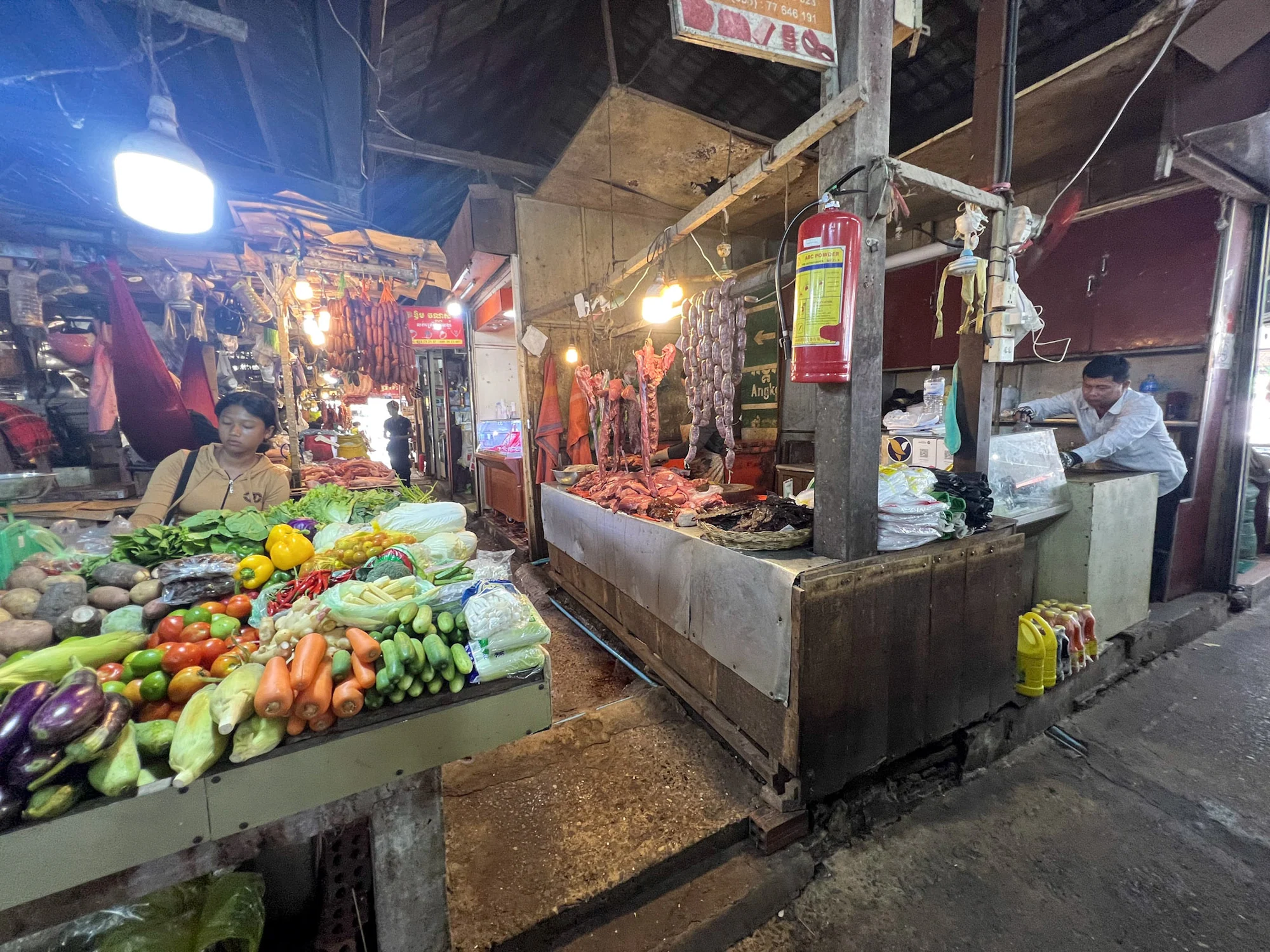 Vegetable and meat stalls lit by bare bulbs inside a covered market.