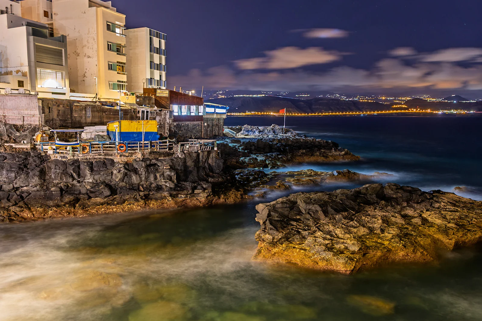 Water washing around coastal rocks beneath buildings at dusk.