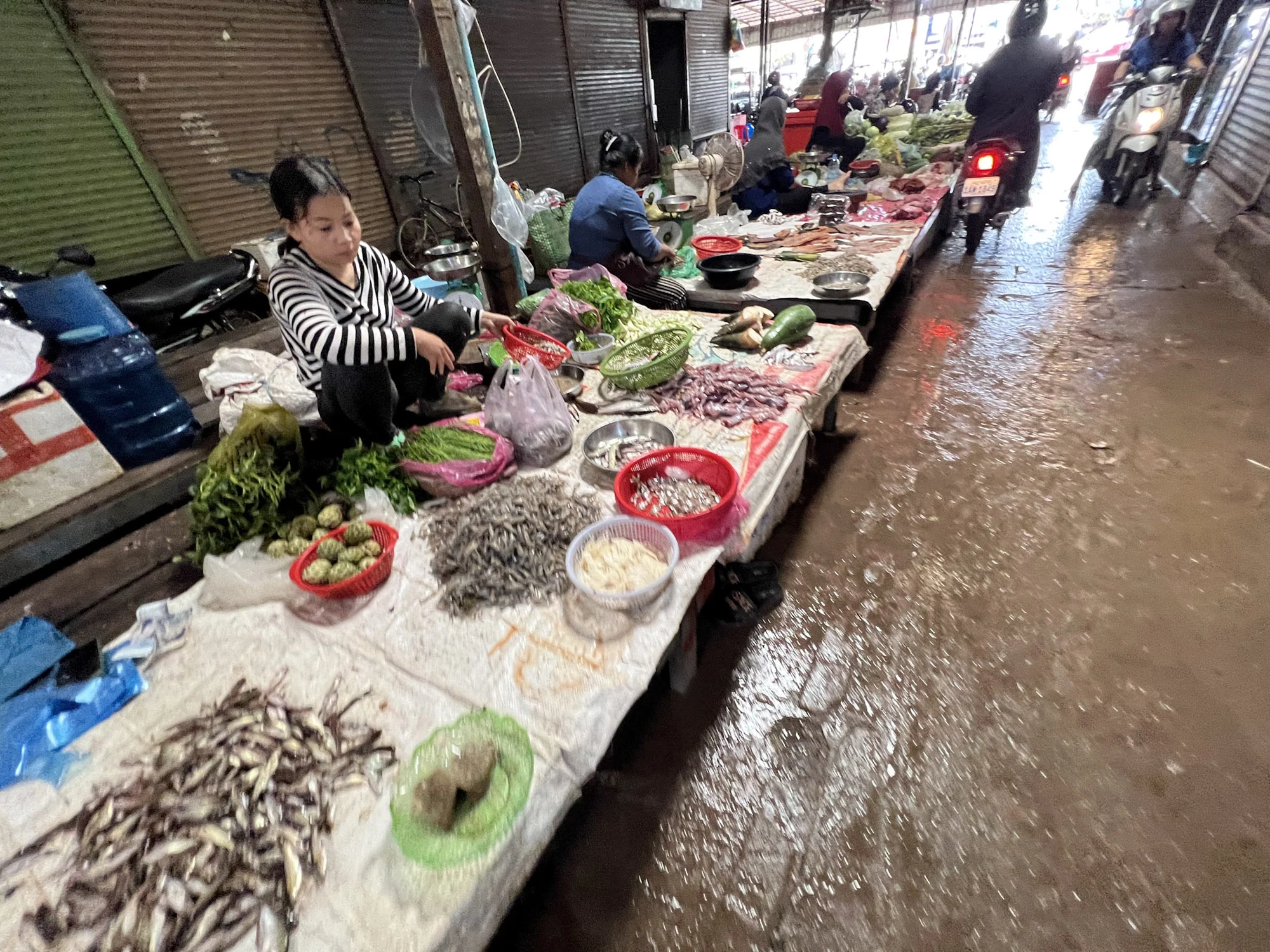 A vendor beside trays of fish and vegetables along a wet market aisle.