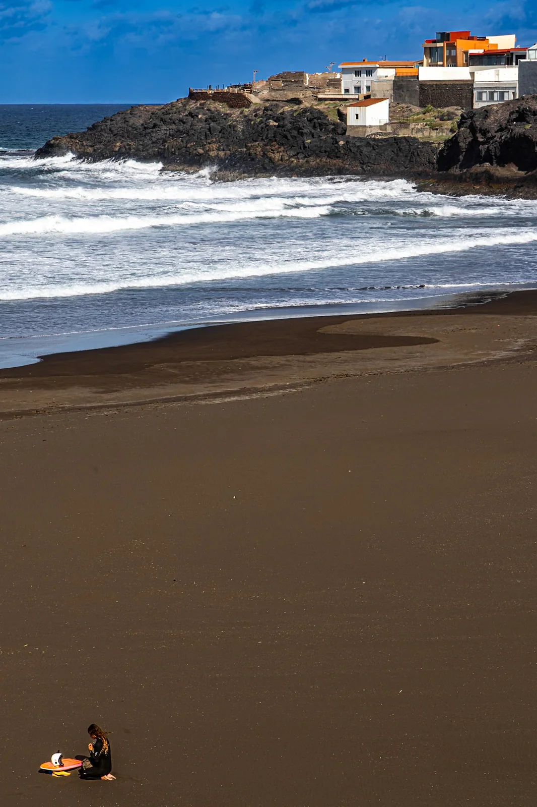 Waves breaking on a dark sand beach, with buildings on a rocky headland.
