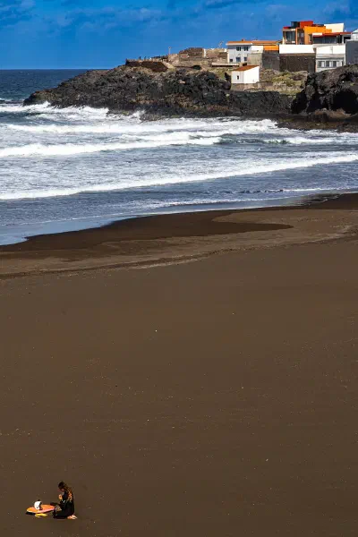 Waves breaking on a dark sand beach, with buildings on a rocky headland.