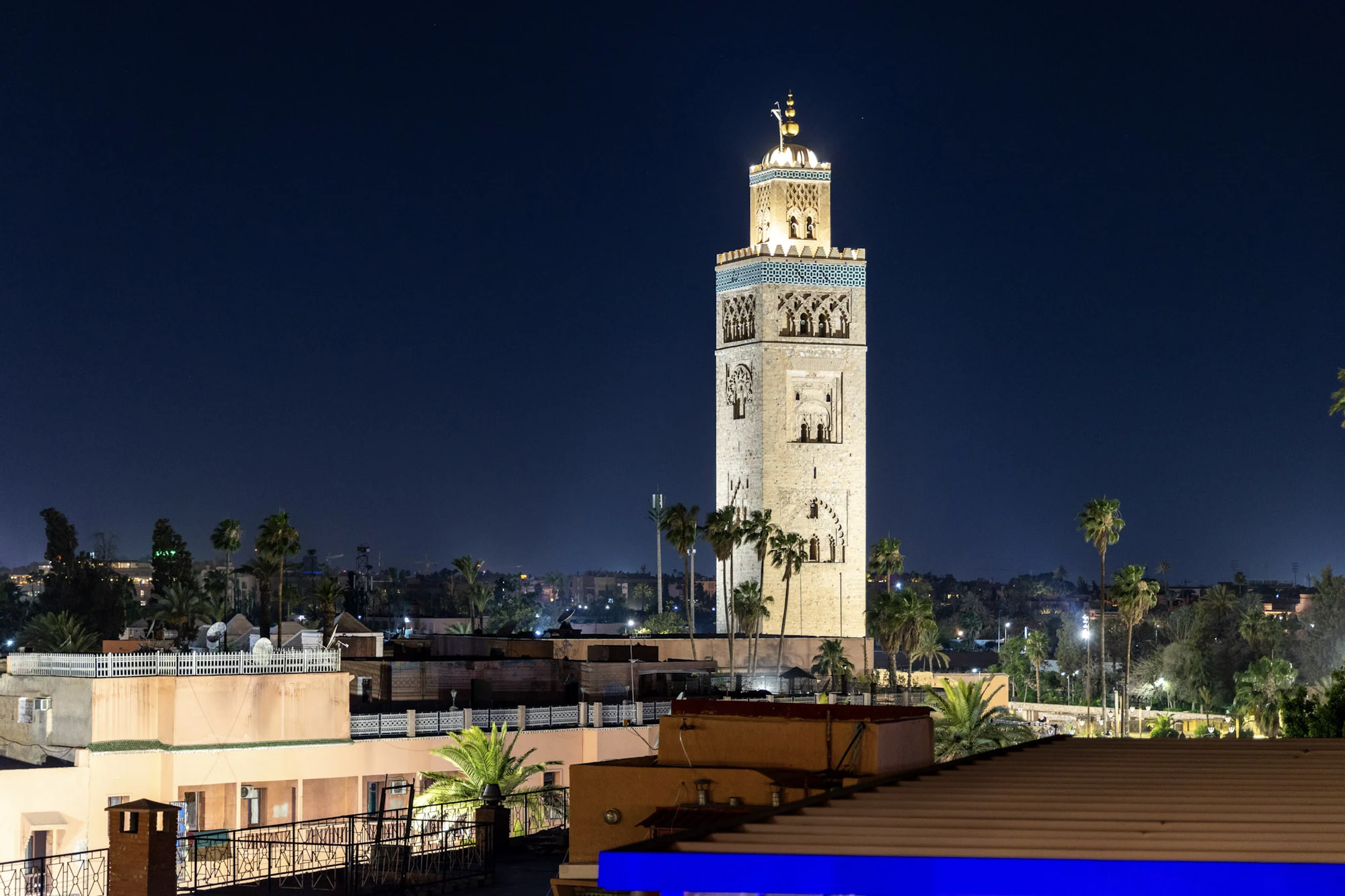 An illuminated minaret above rooftops against a deep blue night sky.