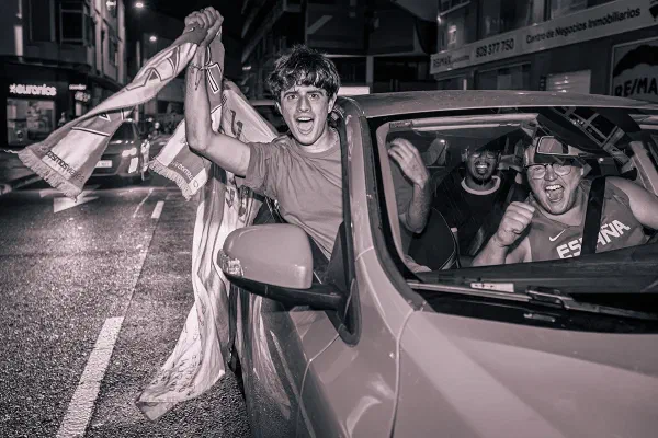 Football supporters celebrating from a car on a city street at night, in black and white.
